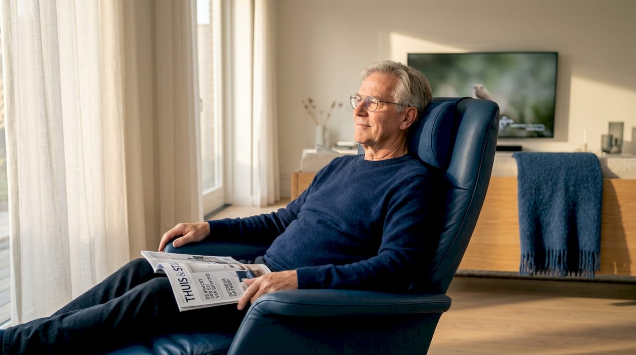Een oudere man zit comfortabel in zijn fauteuil in de woonkamer met een nekkussen om zijn nek.